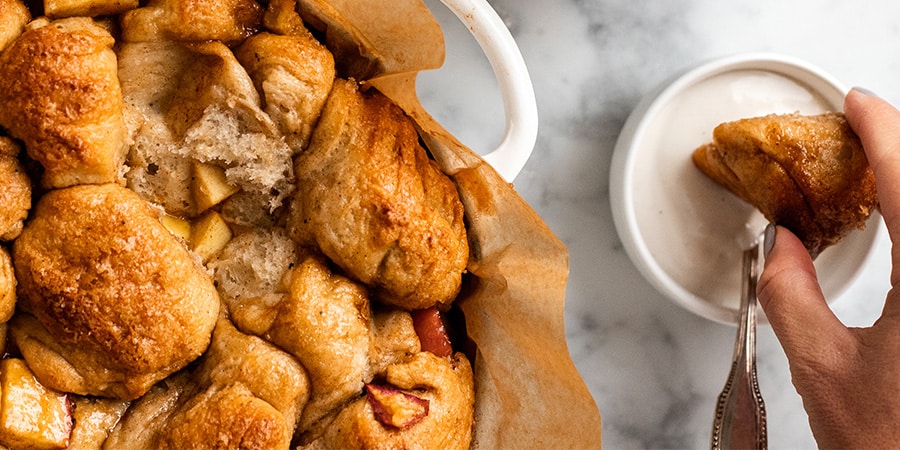A hand dipping a piece of Vegan Monkey bread pulled from a casserole dish into a small bowl filled with Vanilla Cashewmilk Yogurt.