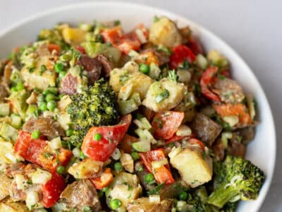 Close up of a large bowl of colorful potato salad that includes bell peppers, broccoli, and peas.
